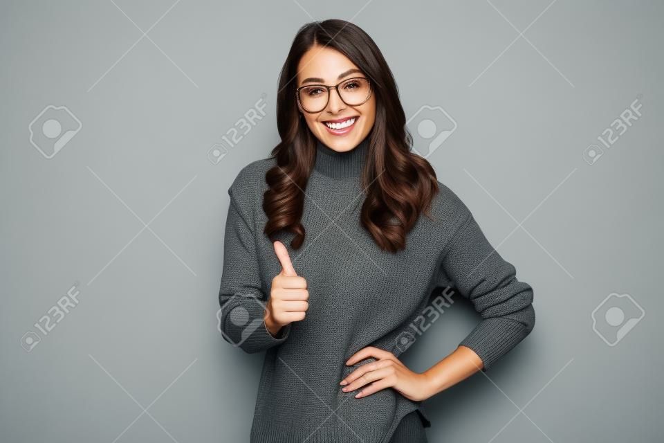 Mulher morena sorridente no suéter e nos óculos com o braço no quadril que mostra o polegar para cima e que olha a câmera sobre o fundo cinzento