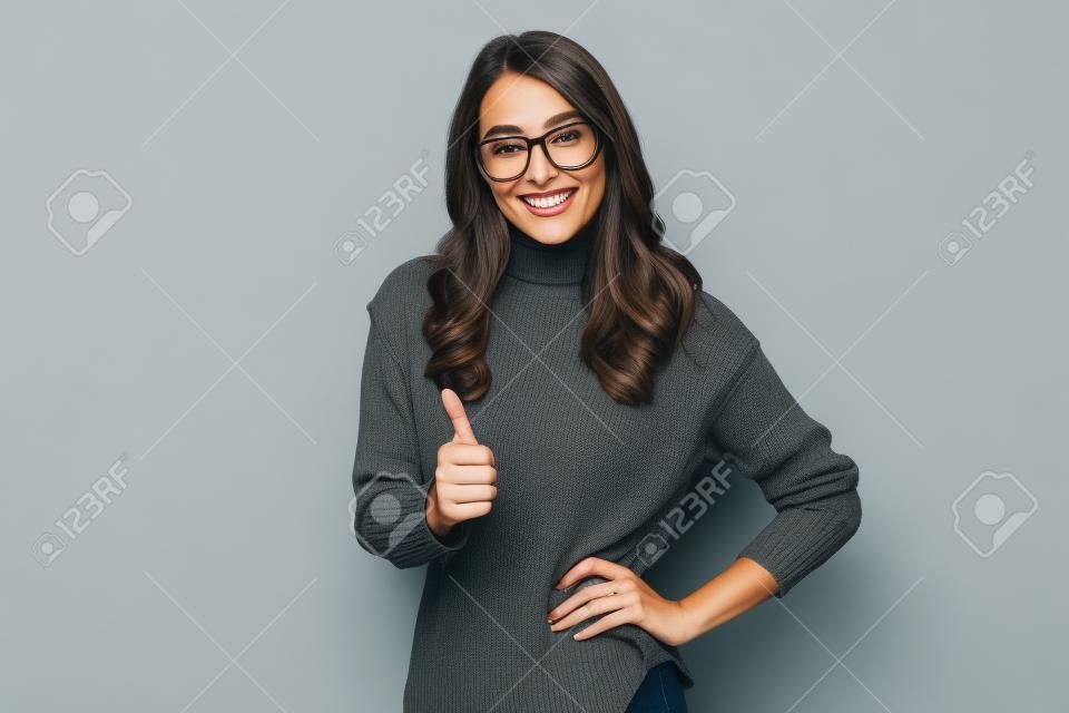 Mulher morena sorridente no suéter e nos óculos com o braço no quadril que mostra o polegar para cima e que olha a câmera sobre o fundo cinzento