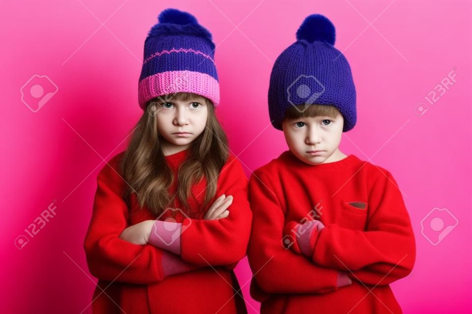 Photo of two displeased angry little children isolated over pink background wearing warm hats. Looking camera.