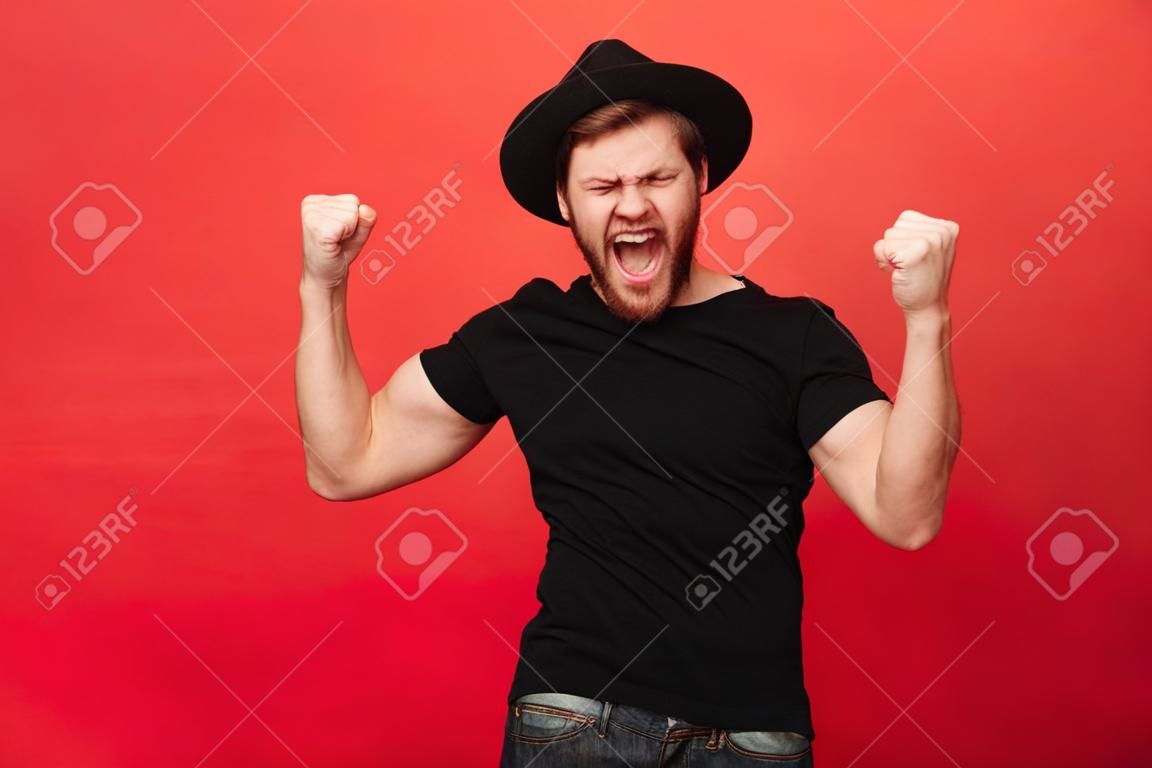 Portrait d'homme macho caucasien extatique en tenue noire se réjouir et serrer les poings dans la joie isolé sur fond rouge