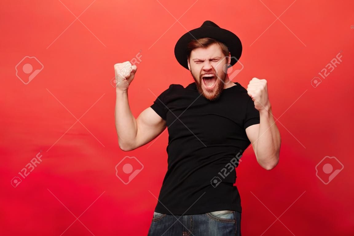 Portrait d'homme macho caucasien extatique en tenue noire se réjouir et serrer les poings dans la joie isolé sur fond rouge