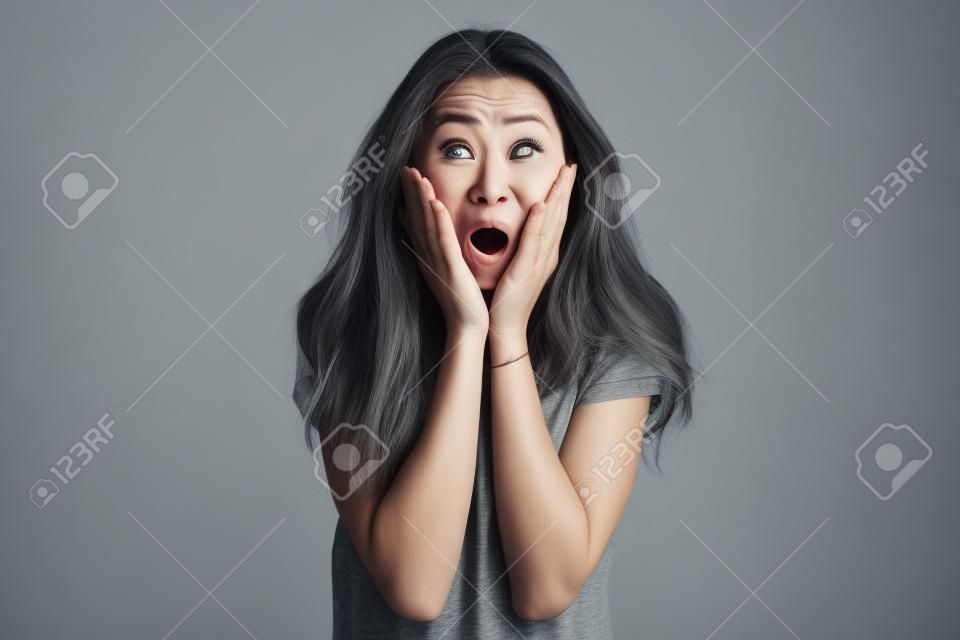 Surprised woman in t-shirt holding her cheeks and looking at the camera with open mouth over grey background