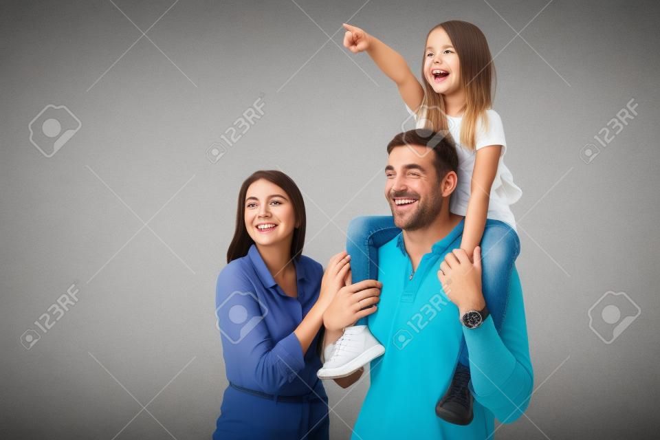 Immagine della donna e dell'uomo della famiglia felice europea che sorride e che osserva da parte mentre la figlia che si siede sul collo di suo padre isolato sopra fondo grigio