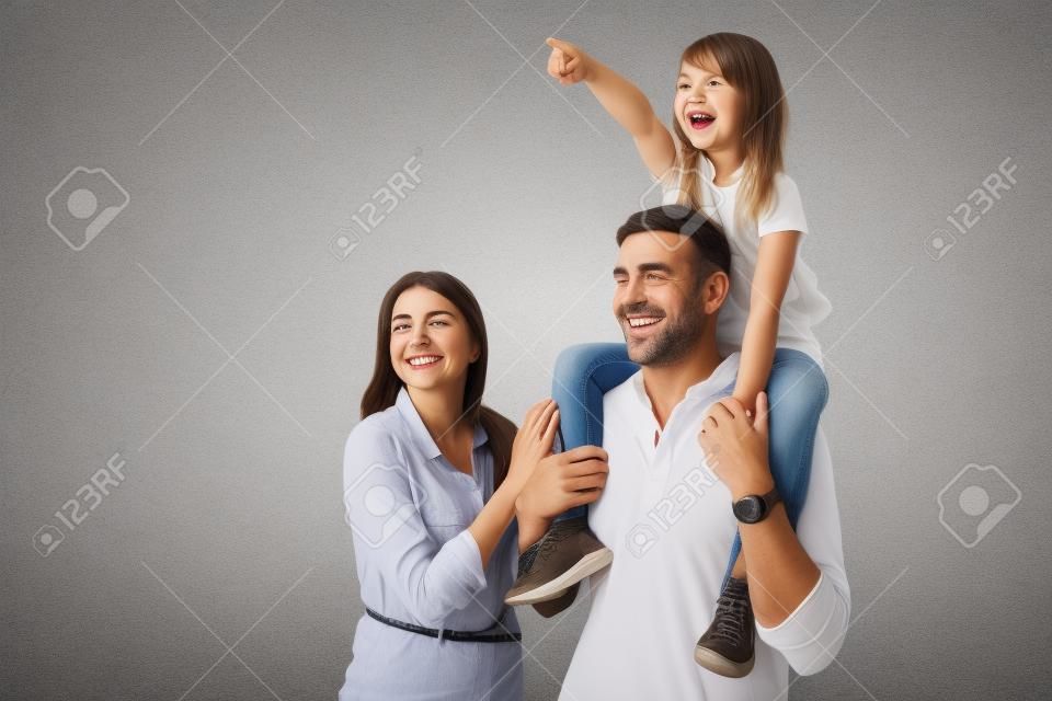 Immagine della donna e dell'uomo della famiglia felice europea che sorride e che osserva da parte mentre la figlia che si siede sul collo di suo padre isolato sopra fondo grigio