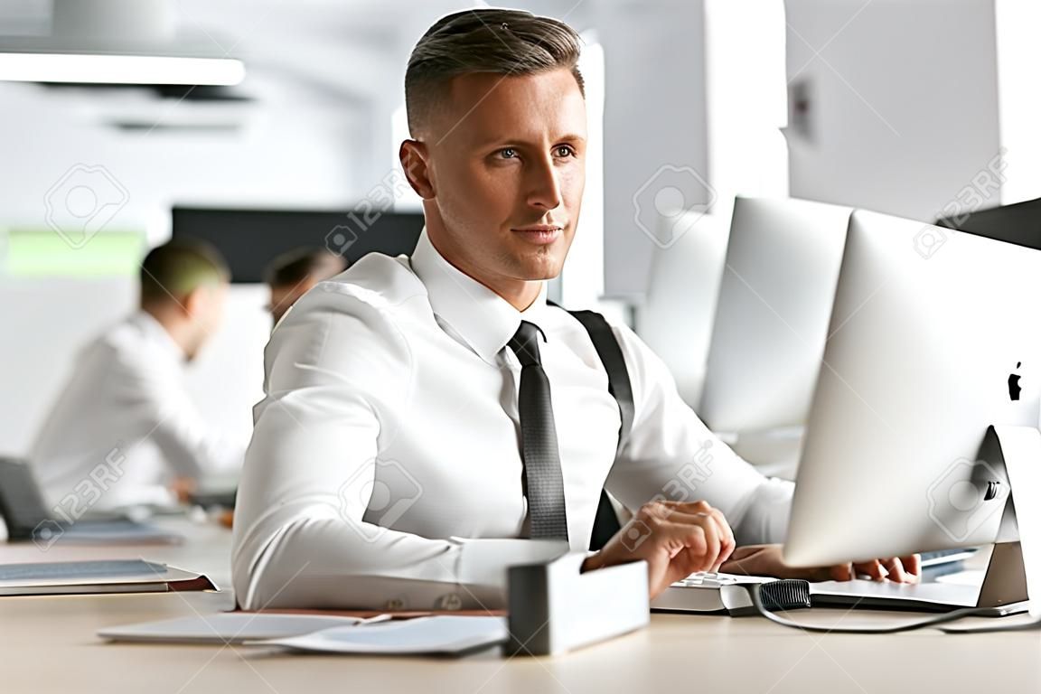 Image of successful employee man 30s wearing white shirt and tie sitting at desk in office and working at computer
