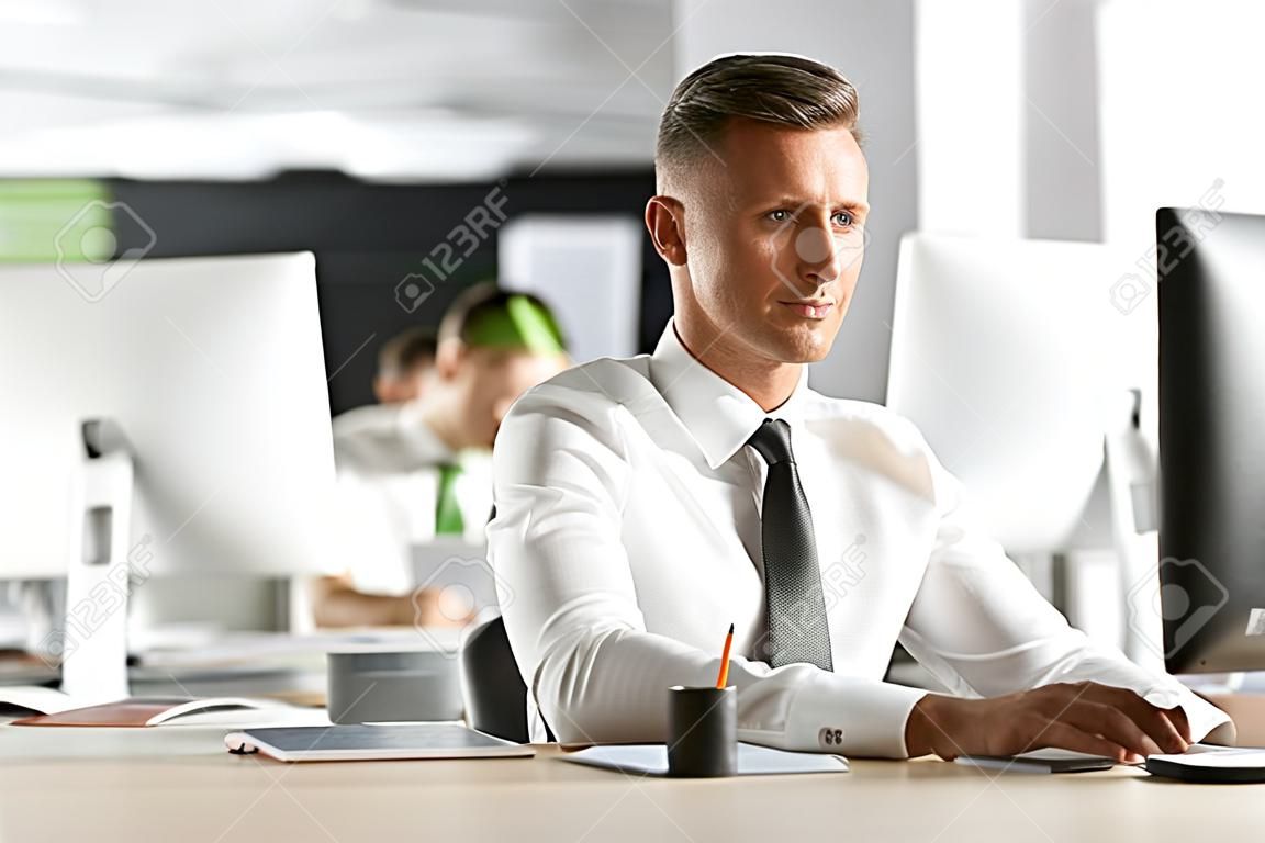 Image of successful employee man 30s wearing white shirt and tie sitting at desk in office and working at computer
