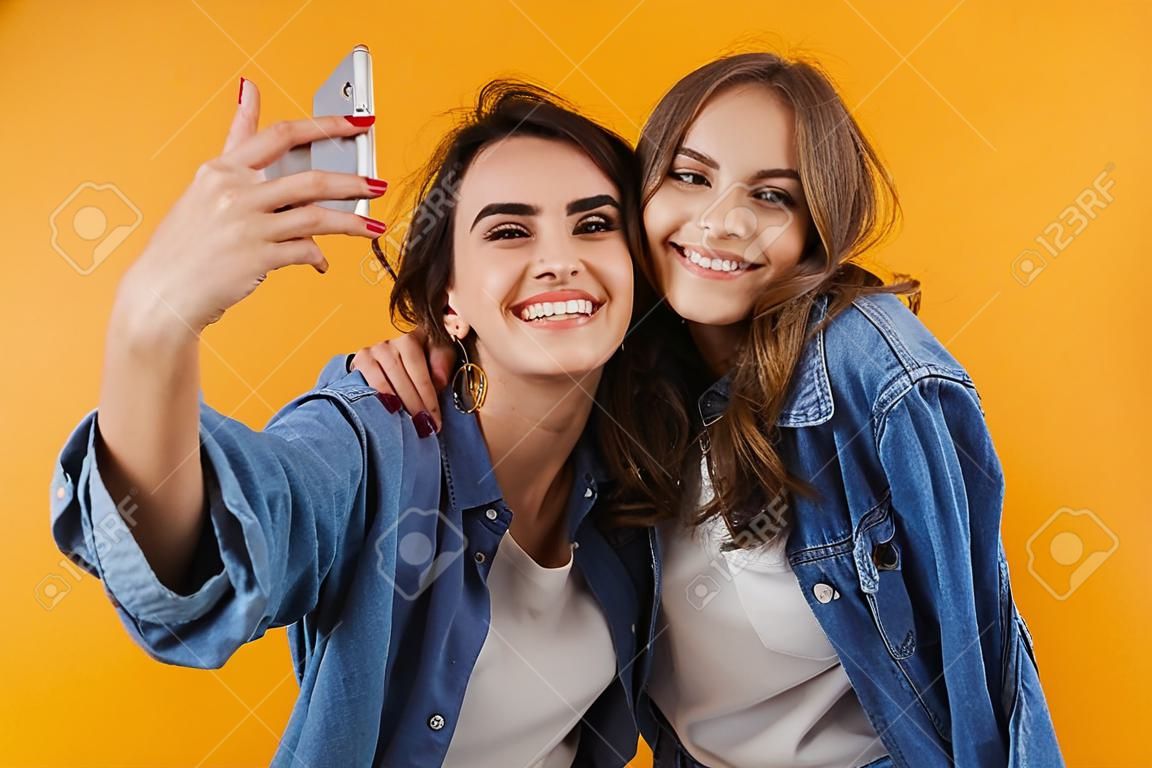 Imagem de feliz animado jovens mulheres amigos isolados sobre fundo de parede amarela tirar uma selfie pela câmera.