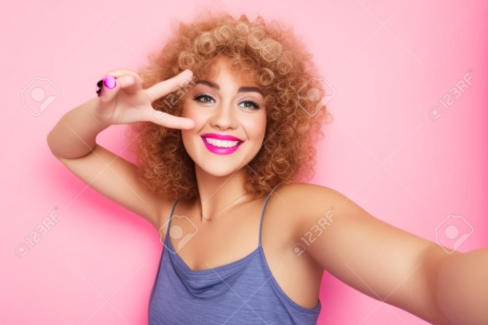Retrato de mujer rizada glamorosa de 20 años sonriendo y tomando una foto selfie aislada sobre fondo rosa