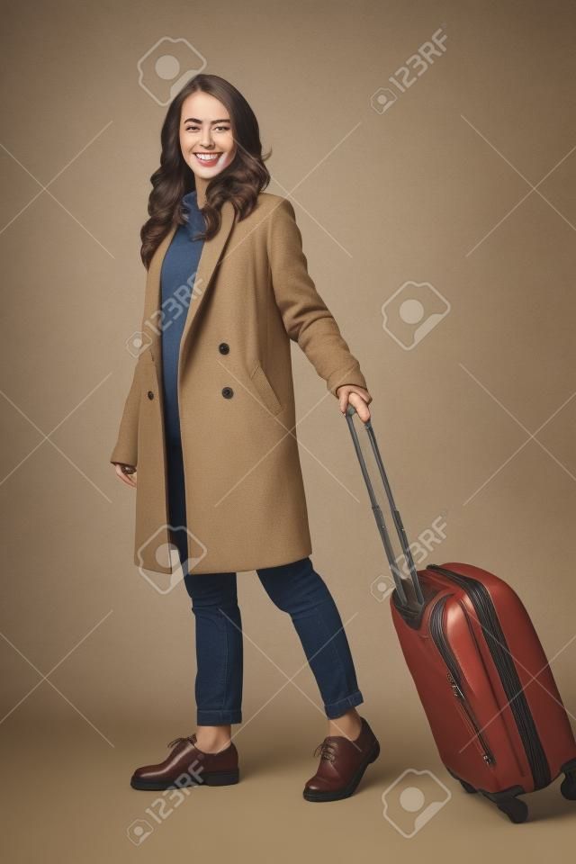 In voller Länge von einer lächelnden jungen Frau, die einen Mantel trägt, der mit einer Reisetasche lokalisiert über beige Hintergrund steht