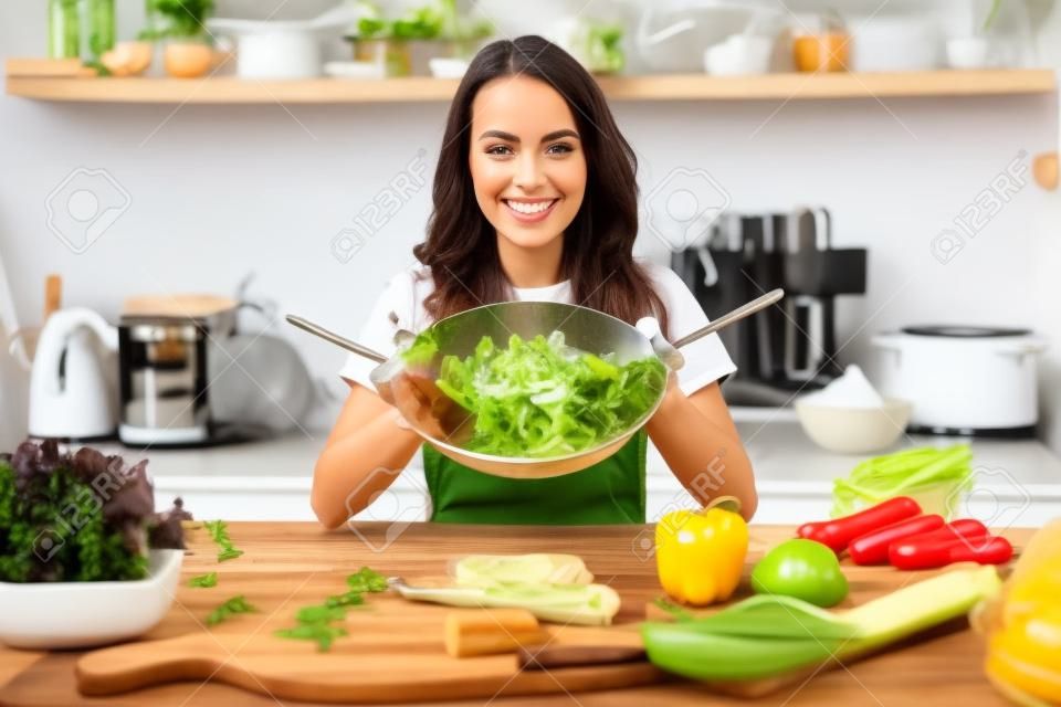 Mulher jovem bonita que veste o avental que cozinha a salada saudável na cozinha em casa, mistura