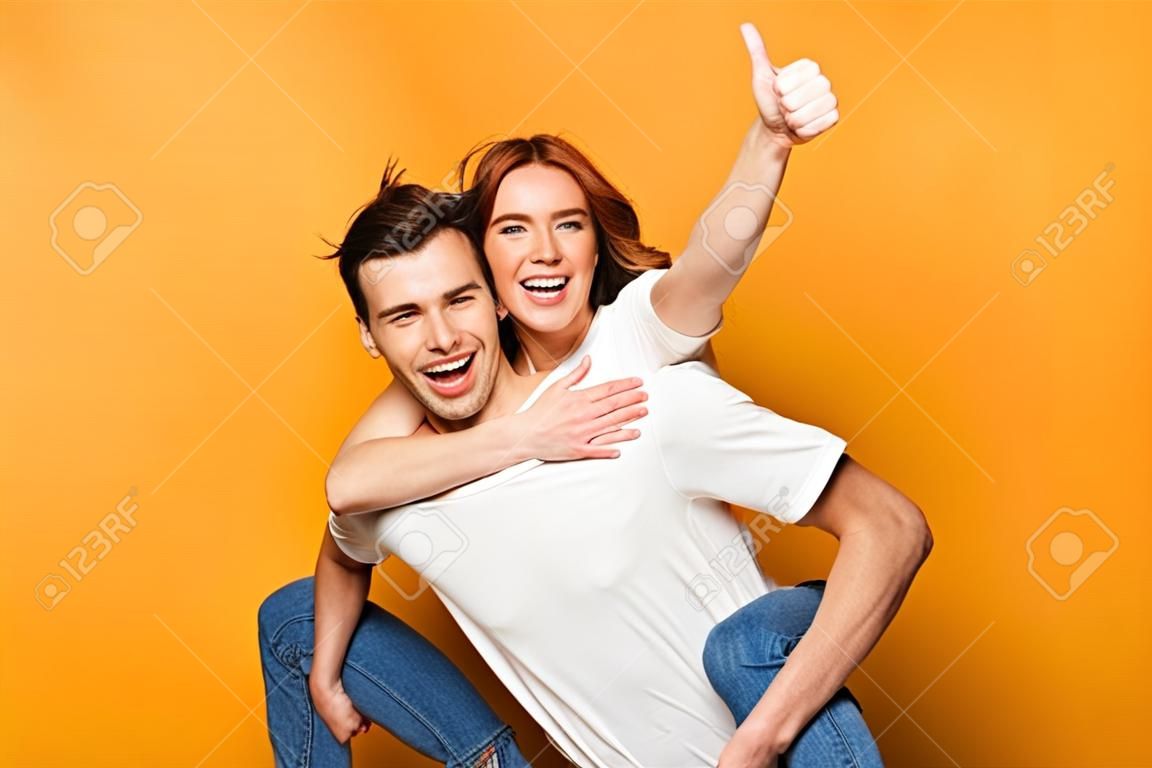 Porträt eines fröhlichen jungen Paares, das isoliert auf gelbem Hintergrund steht, Huckepackfahrt, Daumen hoch thumb
