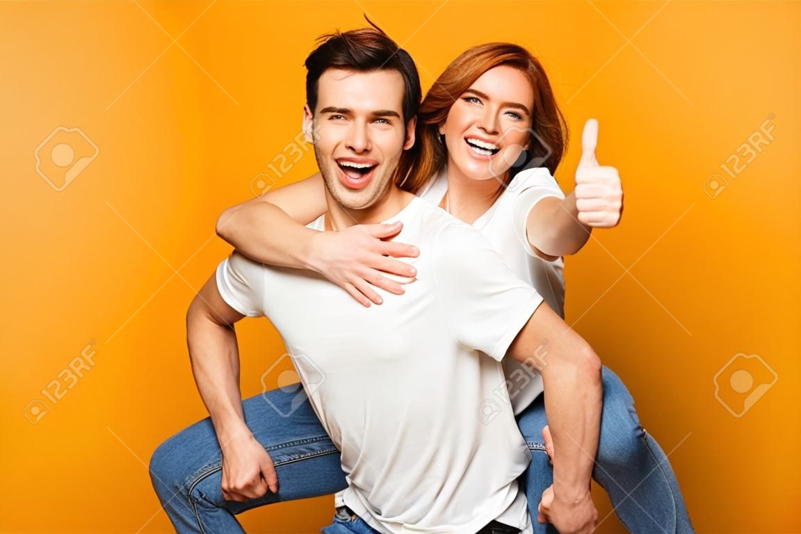 Portrait of a cheerful young couple standing isolated over yellow background, piggyback ride, thumbs up