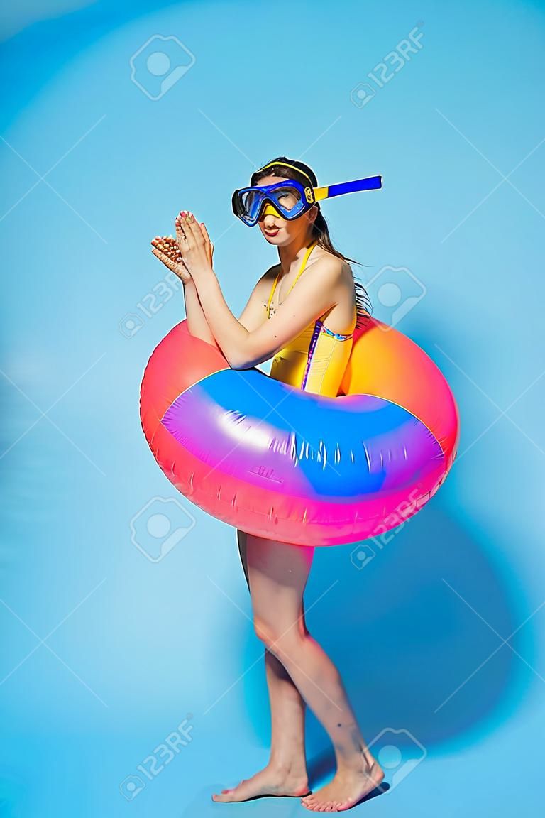 Beautiful young woman wearing swimsuit having fun at the beach with inflatable ring isolated over blue background, wearing flippers and snorkeling mask