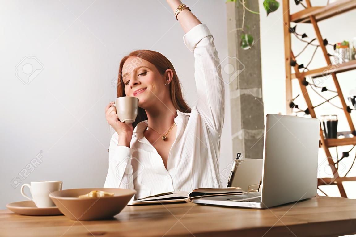 Image of young cute redhead business woman sit indoors in office stretching drinking coffee.