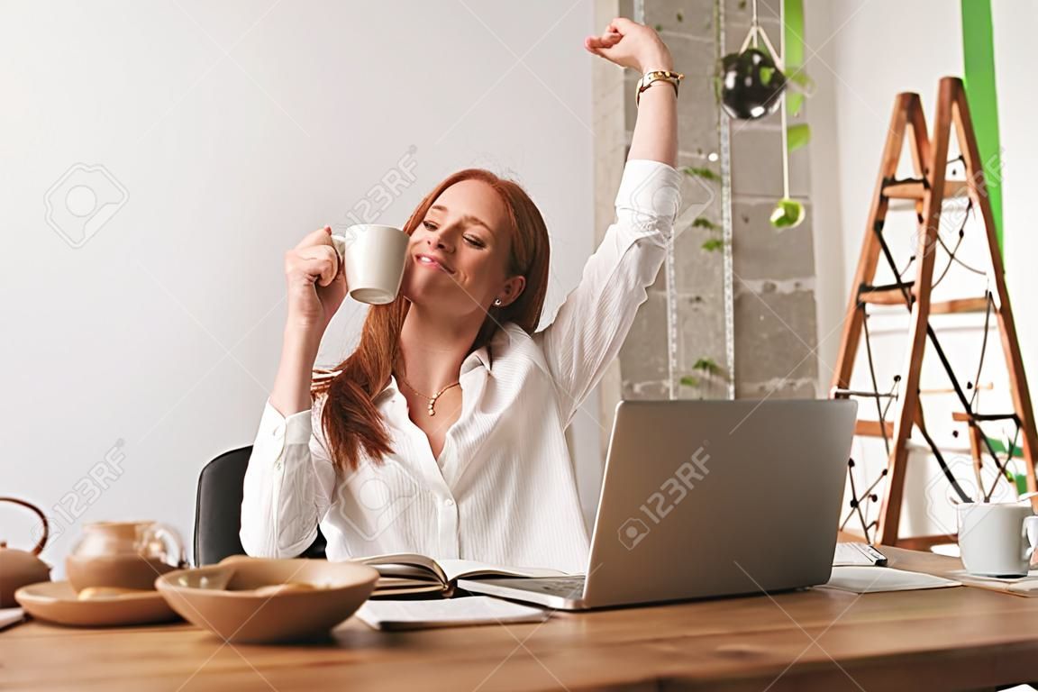 Wizerunek kobiety młody biznes ładny rudy siedzieć w pomieszczeniu w biurze rozciąganie picia kawy.