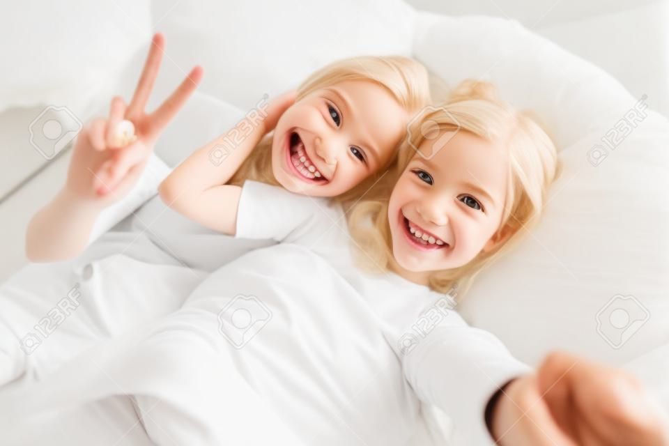 Photo of blonde cheerful mother with daughter gesturing peace sign and taking selfie while lying in white bad