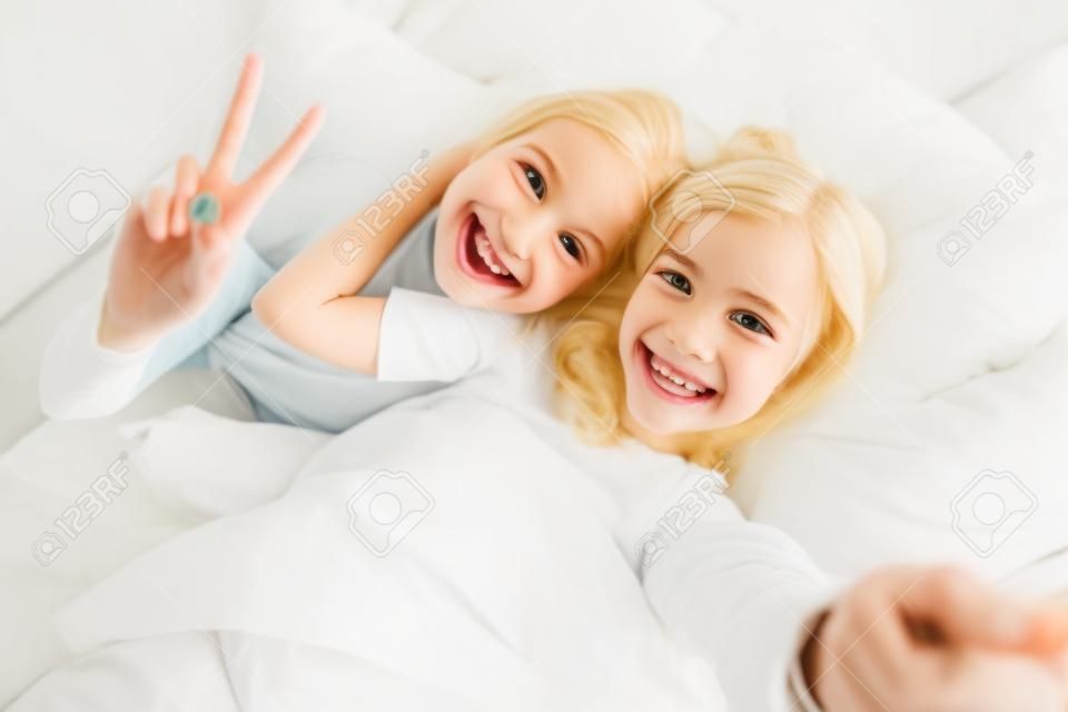Photo of blonde cheerful mother with daughter gesturing peace sign and taking selfie while lying in white bad