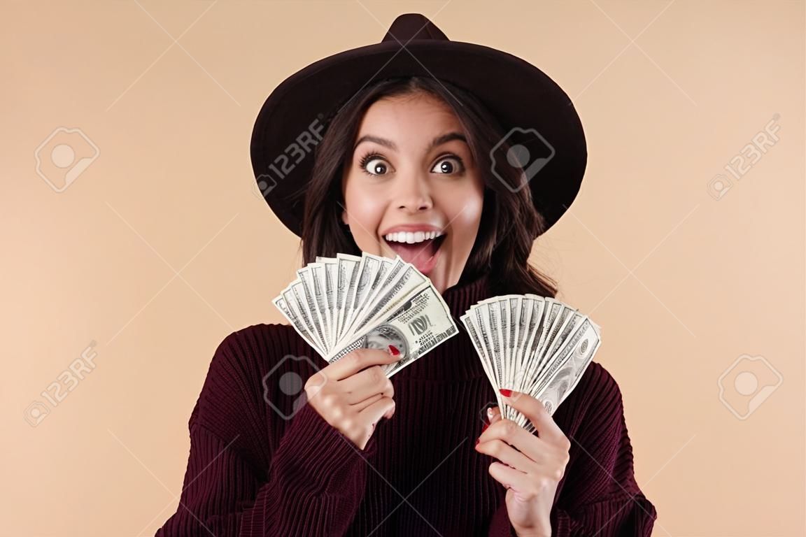 Photo of young happy brunette woman isolated over beige wall background holding money make winner gesture.