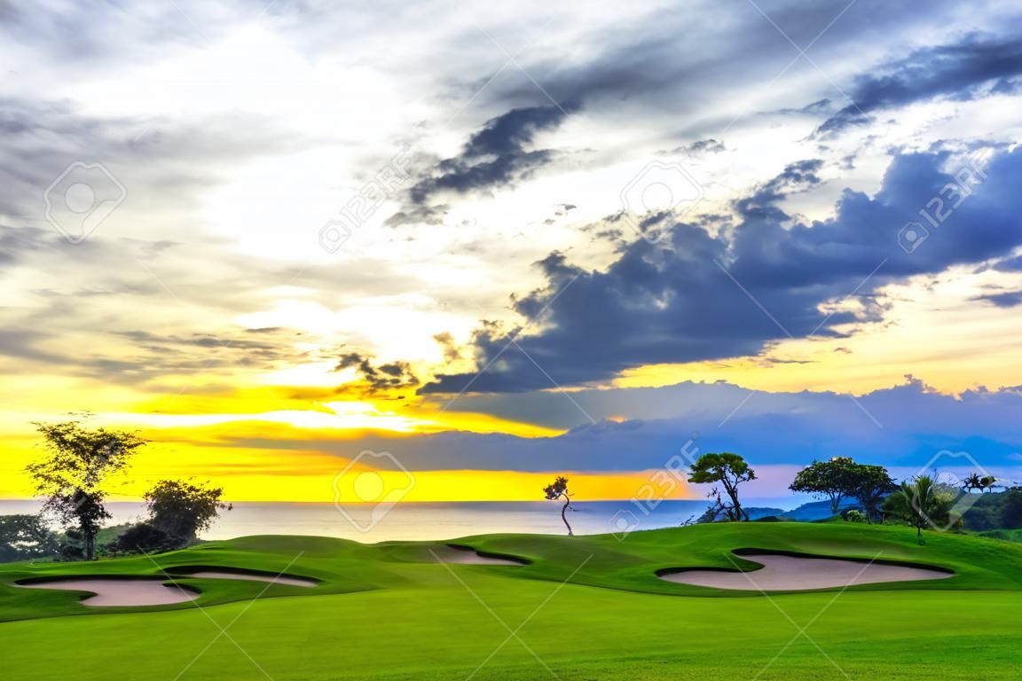 A picturesque green golf course on the background of the sea and the blue sky, in the clouds of which shines a sunny orange light. Sunset on Jimbaran, South Kuta, Bali, Indonesia.
