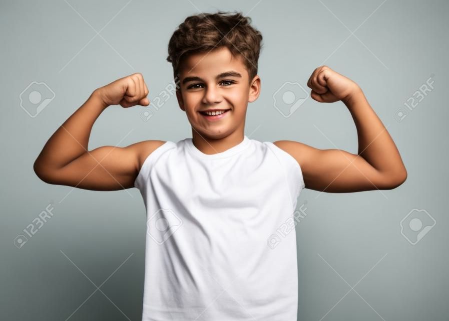 Portrait of funny teen boy raised his hands and shows biceps, isolated on white background. Handsome teenage young boy shows biceps. Happy strong child flexing biceps and looking at camera.