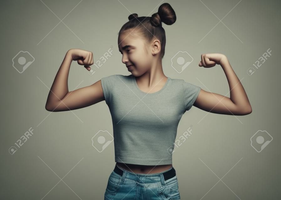 Portrait of funny teen girl raised her hands and shows biceps, isolated on white background. Sad serious cute teenage young girl shows biceps. Upset child flexing biceps.