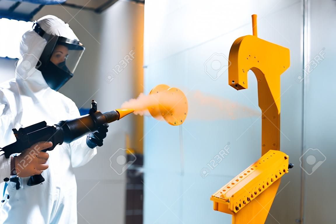 Powder coating of metal parts. A woman in a protective suit sprays powder paint from a gun on metal products
