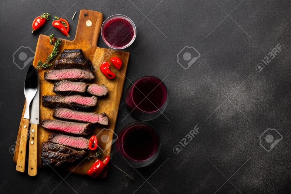 Sliced steak Striploin, grilled with pepper, garlic, salt and thyme served on a wooden chopping Board with a glass of red wine on a dark stone background. Top view with copy space.