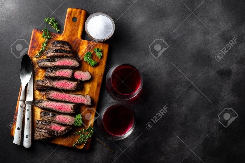 Sliced steak Striploin, grilled with pepper, garlic, salt and thyme served on a wooden chopping Board with a glass of red wine on a dark stone background. Top view with copy space.