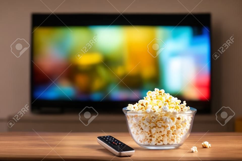 Een glazen kom van popcorn en afstandsbediening op de achtergrond de tv werkt. Avond gezellig kijken naar een film of tv-serie thuis.