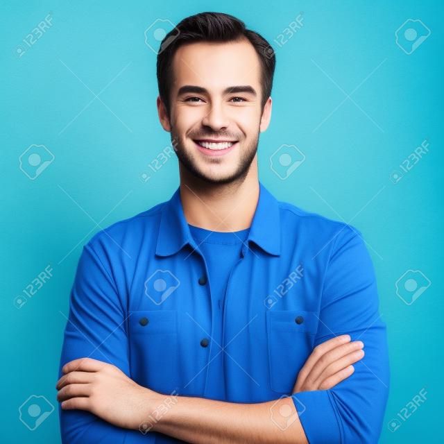 Man portrait. Young happy man with smiling face. Male model in crossed arms pose. Blue background. Guy in casual fashion clothing, studio photo. Square composition picture.