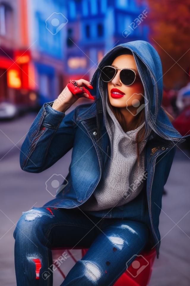 Koncepcja mody ulicznej portret pięknej dziewczyny siedzącej na zewnątrz, niebieskie dżinsy, okulary przeciwsłoneczne, twarde światła, czerwona kurtka, jesienna pogoda