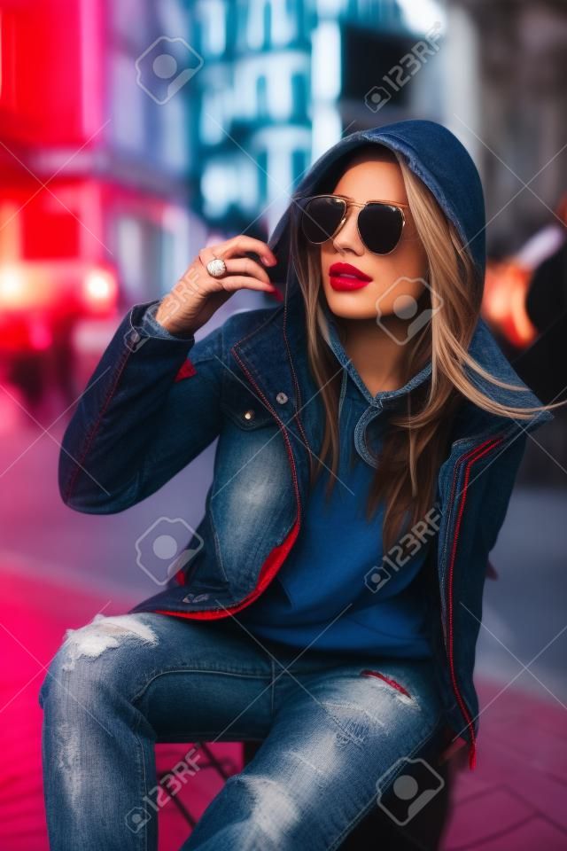 Koncepcja mody ulicznej portret pięknej dziewczyny siedzącej na zewnątrz, niebieskie dżinsy, okulary przeciwsłoneczne, twarde światła, czerwona kurtka, jesienna pogoda