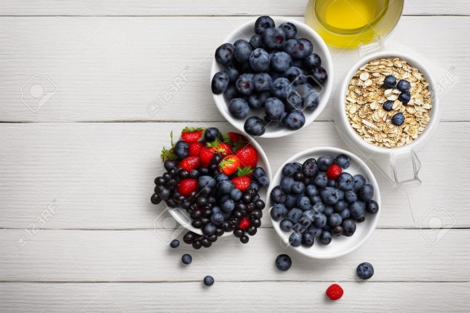 Desayuno del verano. Ingredientes para el desayuno saludable - bayas, fruta y muesli en la mesa de madera blanca, primer plano vista superior horizontal. Tiro macro enfoque selectivo