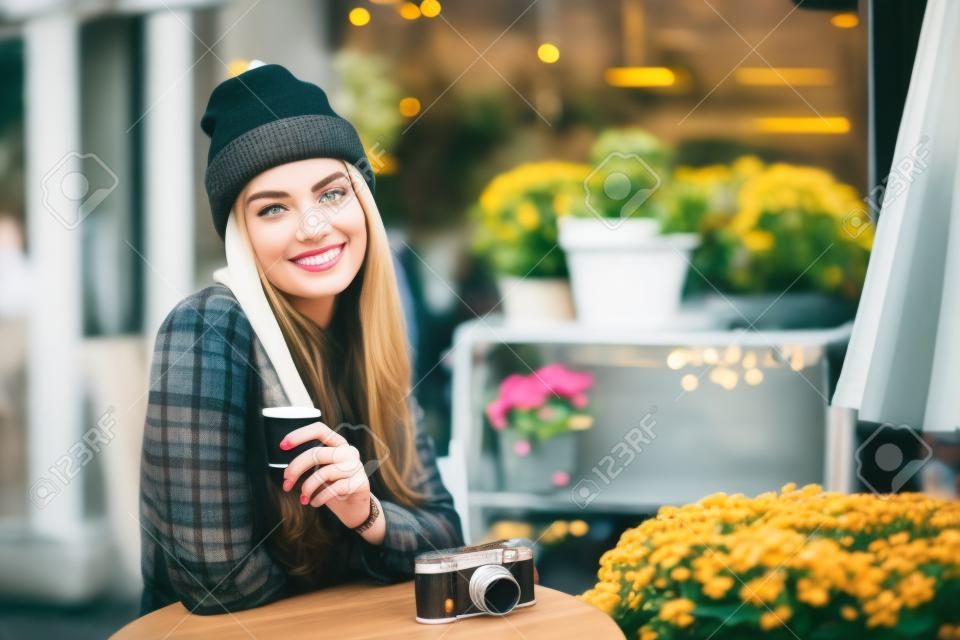 Retrato da bela mulher jovem elegante sentada no café de rua e bebendo café. Hipster com câmera retro velha. Luzes e fundo de flores. Estilo de vida da cidade. Tonificado.
