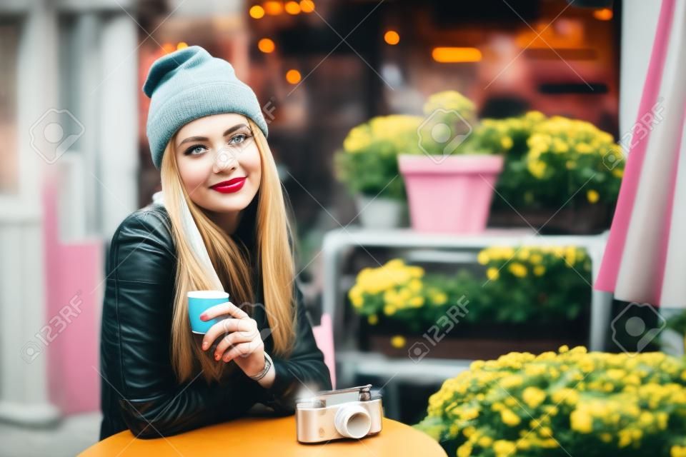 Retrato da bela mulher jovem elegante sentada no café de rua e bebendo café. Hipster com câmera retro velha. Luzes e fundo de flores. Estilo de vida da cidade. Tonificado.