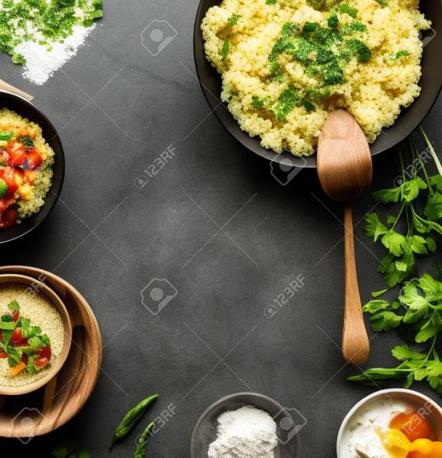 Food background frame with healthy vegetarian couscous pot and bowls with ingredients: vegetables, herbs and feta cheese on dark table, top view, flat lay, frame