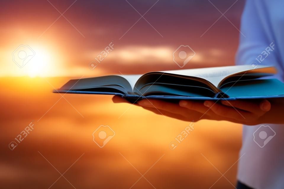 Female hands holding opened hardback book, diary with fanned pages on blurred nature landscape background against sunset sky with back light. Copy space, back to school education concept.