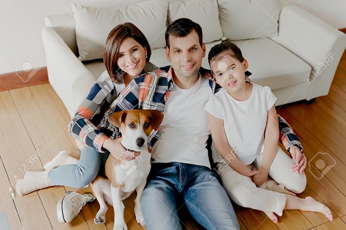 Top view of happy family members pose in spacious room near comfortable sofa, favourite dog poses near, enjoy spare time sit on floor, smile positively. Father, mother and child spend time at home