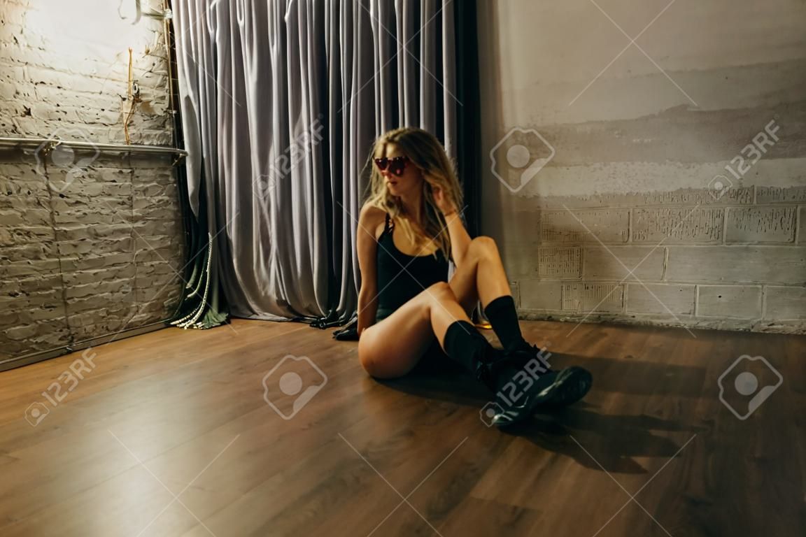 Het schattige blonde meisje draagt in zwart bodysuit en roze zonnebril zitten op de vloer in loft studio. Film foto gain en selectieve focus.