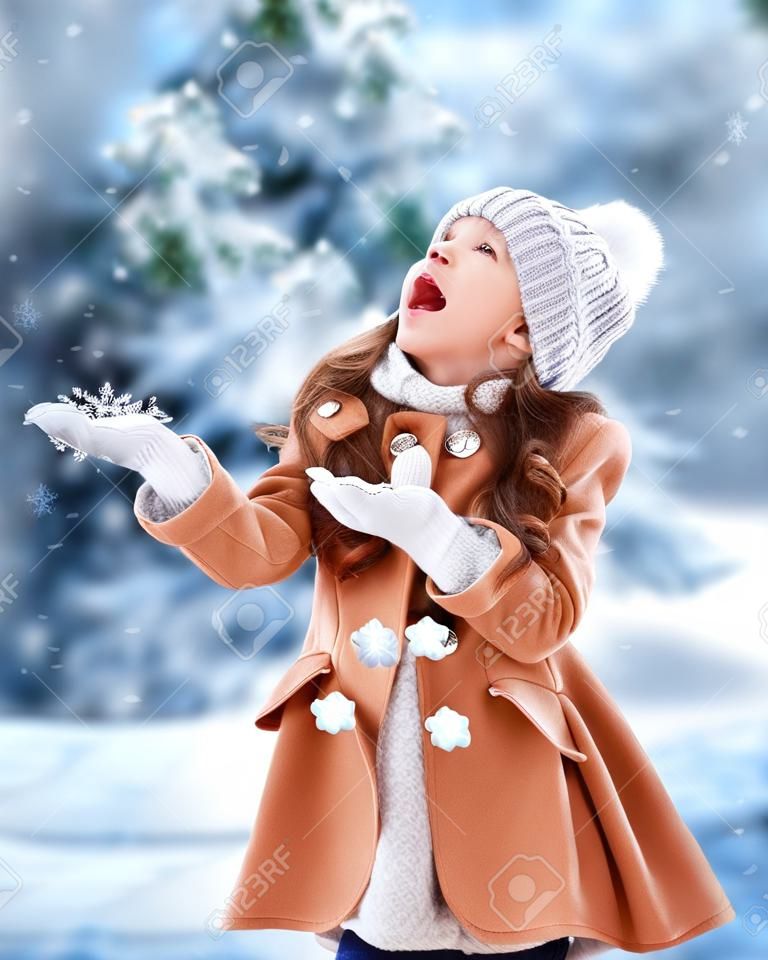 Portret szczęśliwej wesołej dziewczynki zimą w parku łapie płatki śniegu ustami, ciesząc się zimą w parku.