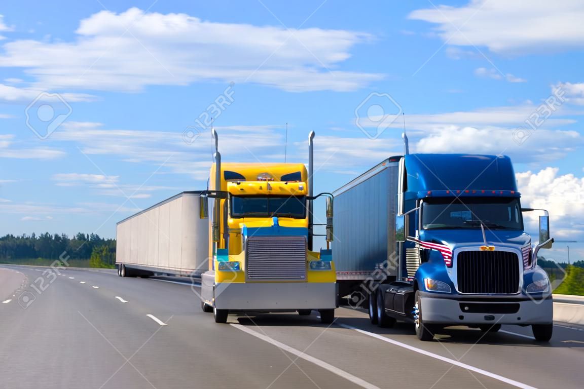 Due camion semi di vari modelli e produttori, un camion semi americano classico giallo con un rimorchio alla rinfusa e un camion semi americano moderno blu con un rimorchio alto per carichi alla rinfusa che corrono lungo un'ampia autostrada uno accanto all'altro