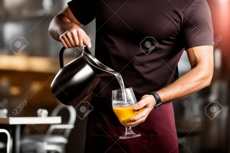 Strong slim man with muscular muscles working as a waiter in a restaurant in a black t-shirt and burgundy apron pours water from a stainless steel jug into a customers glass