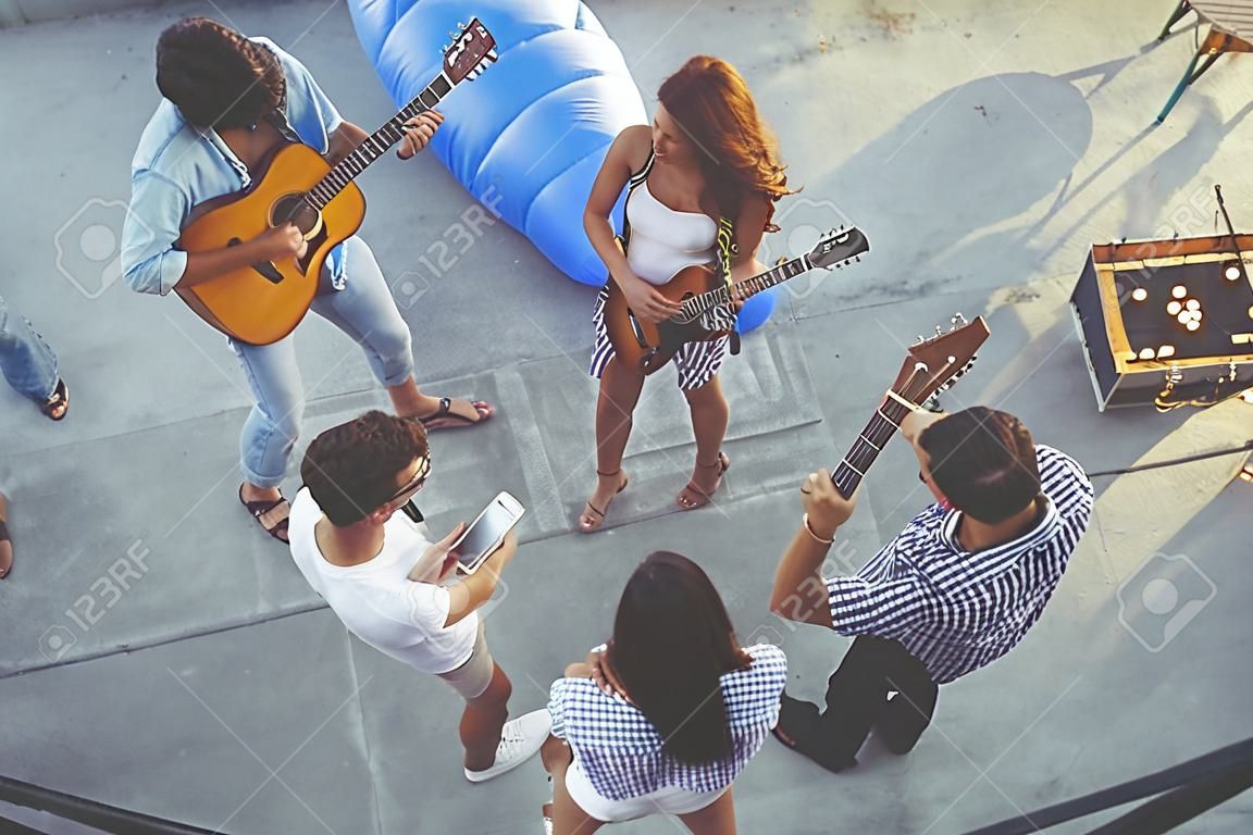 Top view of a group of young friends having fun at a rooftop party, playing the guitar, singing and enjoying hot summer days. Focus on the couple in the middle