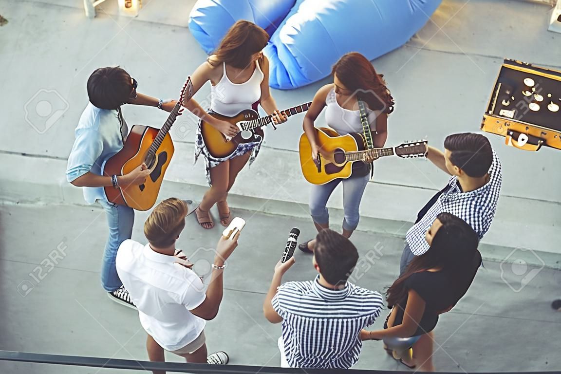 Top view of a group of young friends having fun at a rooftop party, playing the guitar, singing and enjoying hot summer days. Focus on the couple in the middle