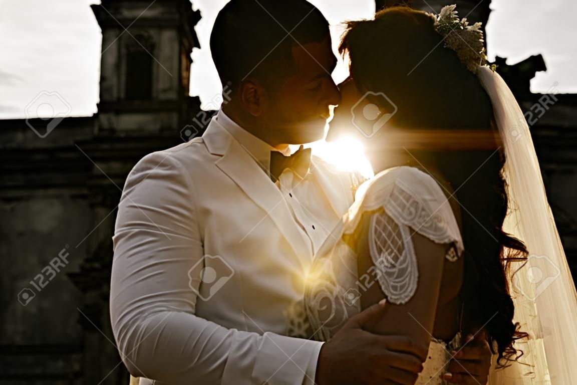 오래된 르네상스 궁전 근처에 서 있는 웨딩 커플의 놀라운 초상화. 그들은 마주보고 서 있습니다. 그들 사이의 햇빛 때문에 우리는 그들의 실루엣만 볼 수 있습니다.