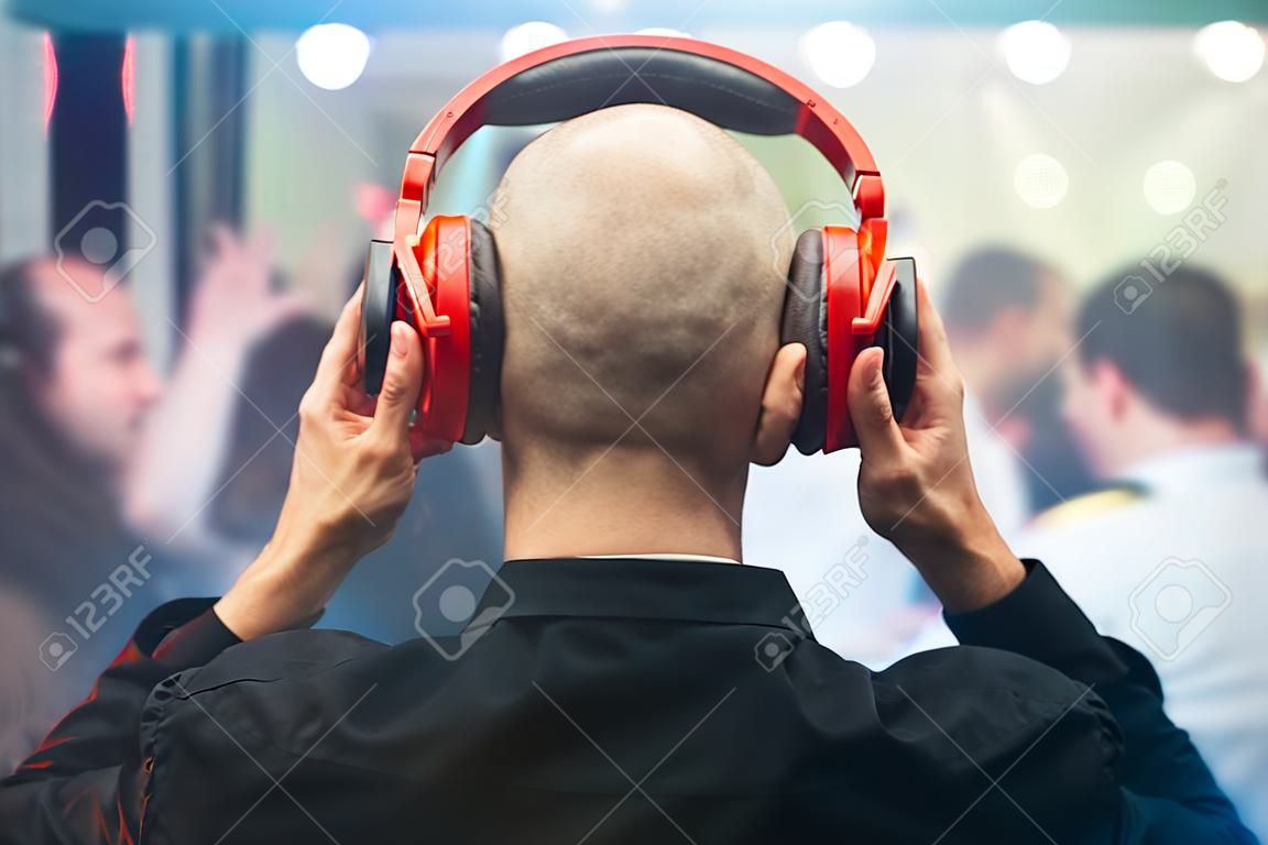 DJ mit roten Kopfhörern bei Nachtclubparty unter dem weißen Licht und Leute drängen sich im Hintergrund. Charismatischer Discjockey am Plattenspieler.