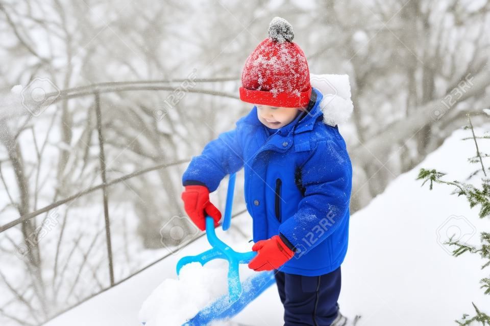 Kleiner Junge macht Schneebälle mit Schneeballhersteller. Glückliches Kind, das mit Schnee spielt. Kaltes Winterwetter. Winteraktivitäten für Kinder