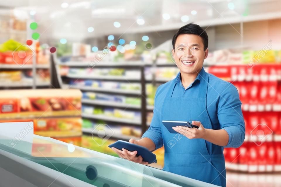 Portrait d'un gérant de magasin asiatique, d'un homme avec une tablette vérifiant la date d'expiration des produits, d'un vendeur souriant et regardant la caméra près d'un réfrigérateur dans un supermarché