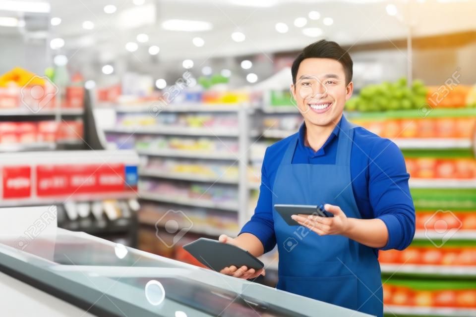 Portrait of Asian store manager, man with tablet checking expiration date of products, seller smiling and looking at camera near refrigerator in supermarket