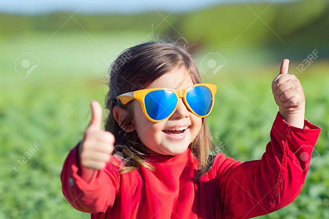Glückliches kleines Mädchen mit großen Sonnenbrillen auf der Wiese zu Fuß und in die Sonne schaut. Mädchen mit den Händen in der Luft machen Daumen nach oben